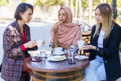 Three women chatting over coffee and baked goods on sunny outside table