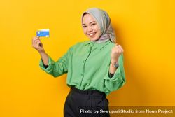 Muslim woman in headscarf and green blouse holding her credit card and clenching fist in celebration 5leLN4