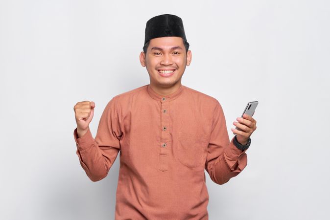 Muslim man in kufi hat holding mobile phone in one hand with other fist clenched