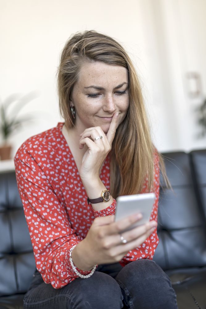 Coy woman at home sitting on couch looking at cell phone
