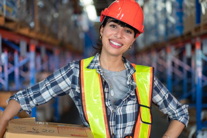 Woman in hard hat smiling and carrying in box in warehouse