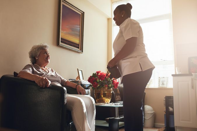 Mature woman sitting on a chair at home with female caregiver standing by