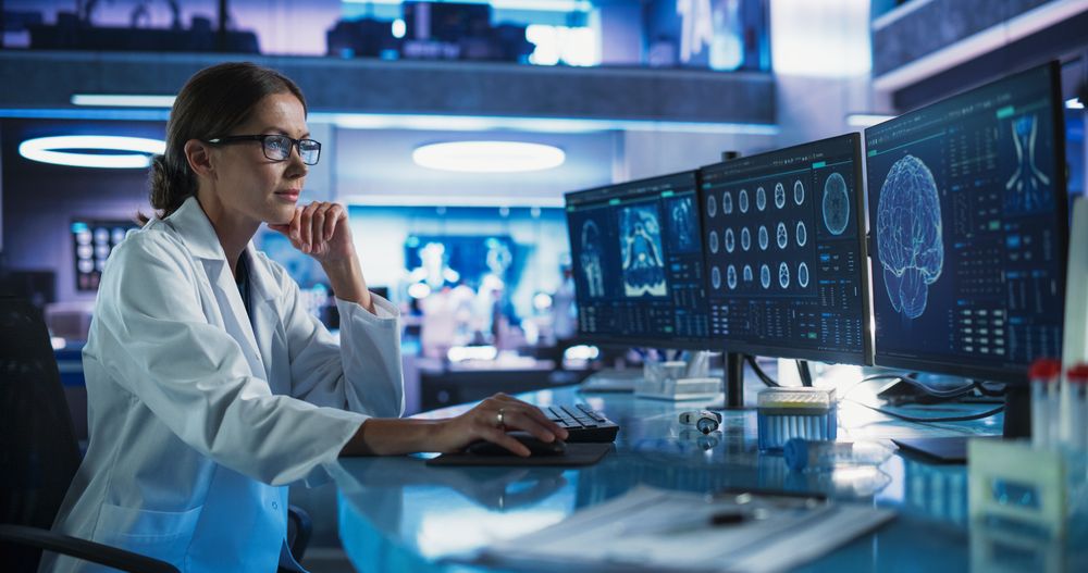 Female Neuroscientist Using Desktop Computer To Analyze MRI Scans Of Human Brain In Busy Research Center Laboratory.