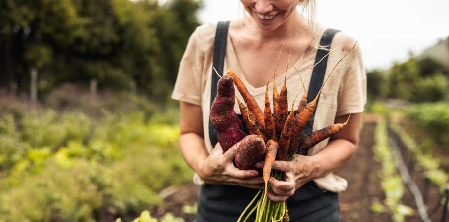 Cheerful female farmer holding freshly picked carrots and sweet potatoes on her farm