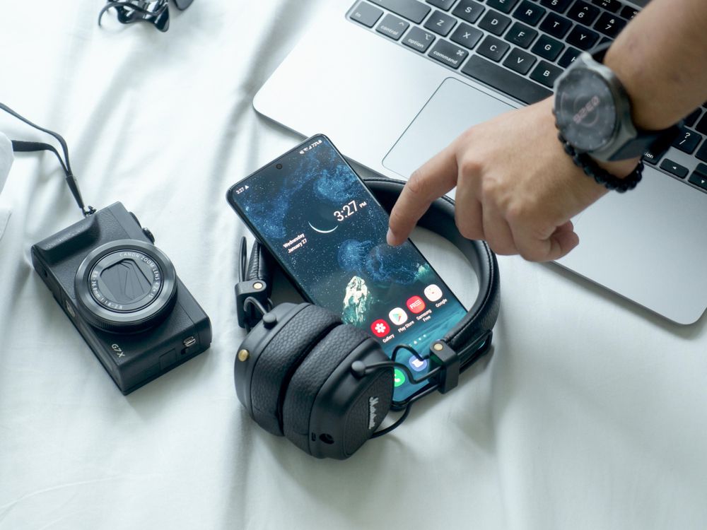 Cropped image of a hand about to touch smartphone on a desk beside laptop and a camera and headphones