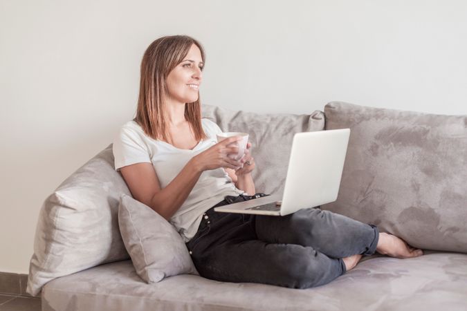 Woman relaxing on sofa with a cup of coffee working on her laptop in the morning