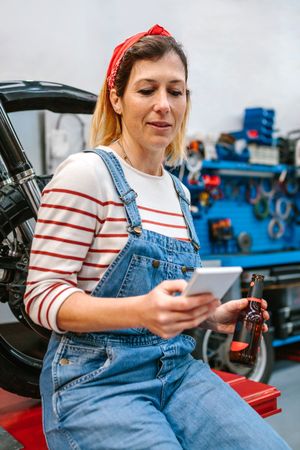 Mechanic woman looking phone and holding beer sitting in front of motorcycle on factory