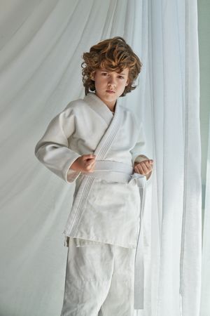 Confident preteen kid in kimono practicing karate near window