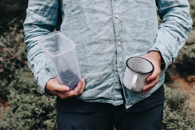 Man in blue linen shirt holding tin cup and plastic container with blueberries