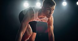Strong Young Man Starting a Race From Track Starting Blocks Position on a Dark Stadium in the Evening. Portrait of a Fit Male Sprint Runner Participating for an International Competition
