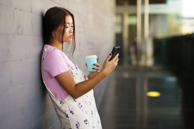 Asian female wearing casual clothes taking selfie with phone while leaning against wall with coffee