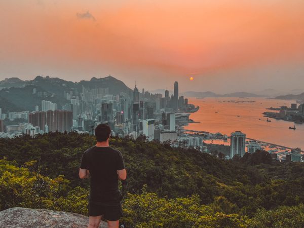 Back view of man standing on top of mountain looking at city buildings during sunset