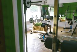 Looking through gym equipment at man in green t-shirt lifting heavy bar exercising arms and chest