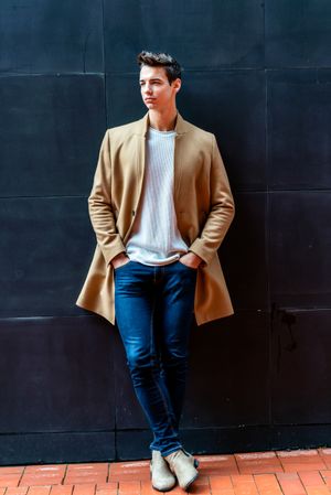 Young man leaning on dark wall outside looking away from camera 