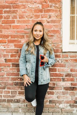 Woman in denim jacket and dark pants leaning against red brick wall