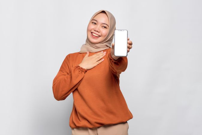 Happy Muslim woman smiling holding up smart phone with hand on chest