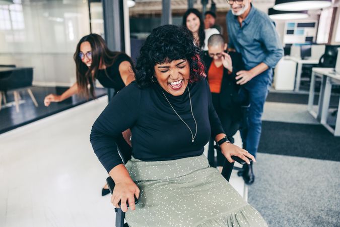 Businesswoman laughing while being pushed in office chair by coworkers