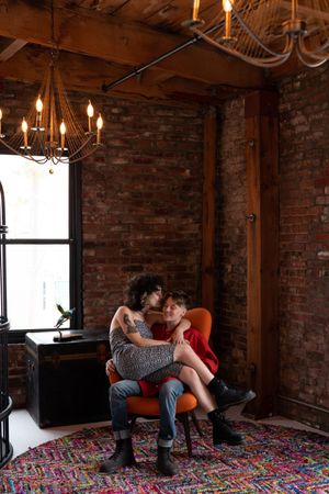 Woman in fashionable jumpsuit sits on her partner’s lap and gives her a kiss