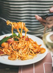 Man in striped shirt eating pasta with fork, wine, and smartphone, close up