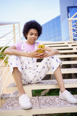 Woman in floral overalls sitting on stairs checking phone, vertical