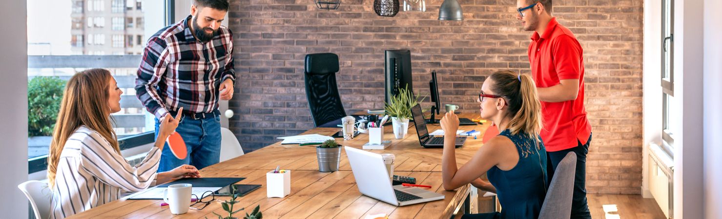 Colleagues talking in office while two of them having fun playing ping pong over coworking table