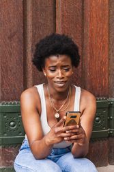 Smiling Black woman using smartphone sitting near building