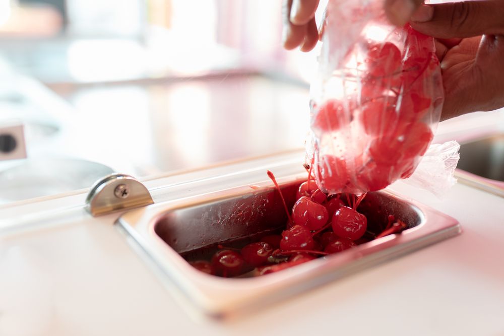 An ice-cream parlor worker is dropping some cherry toppings into the counter tray
