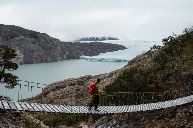 Person with backpack walking on suspension bridge near body of water and mountains