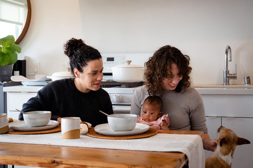 Two women with newborn baby petting their dog at their dining table