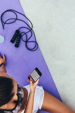 Top view of young woman sitting on exercise mat using fitness app on her mobile phone