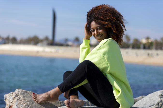 Carefree female sitting on rocks near shoreline in bright green shirt