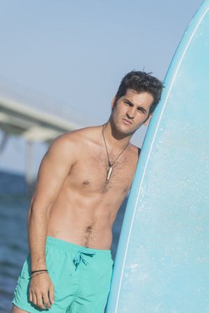 Front of shirtless male surfer standing next to blue board on the coast, vertical composition