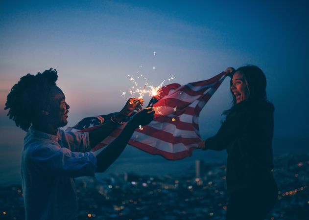 A happy woman holds the America flag in the wind while a man holds sparklers at dusk