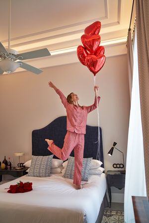 Happy woman with red heart shaped balloons jumping on bed