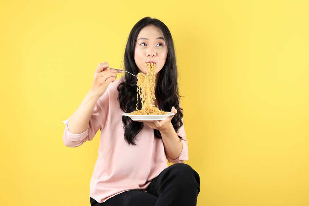 Happy Young Asian Woman Sitting on a Chair Wearing Pink Shirt, Eating Noodles on White Plate using Fork.