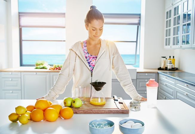 Fit woman in zip up hoodie making fresh fruit juice in kitchen