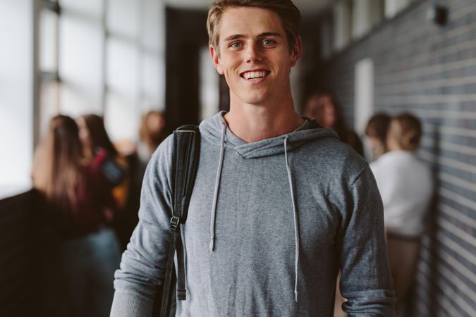 Portrait of happy young guy with bag walking in university corridor with classmates in background