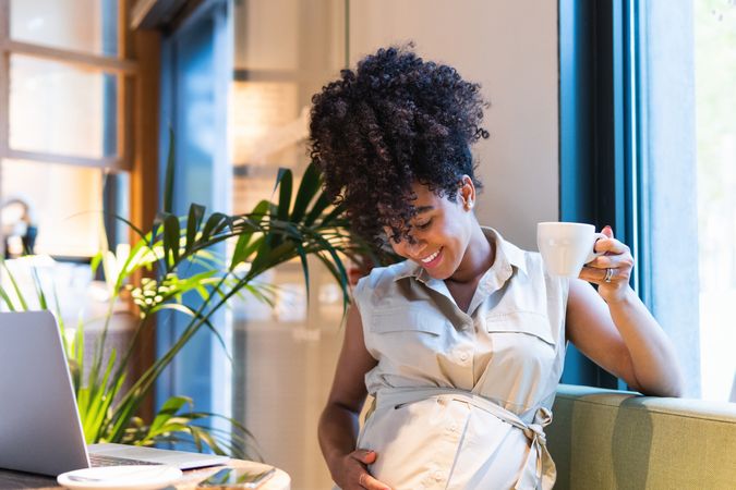 Smiling Black woman drinking coffee and touching tummy