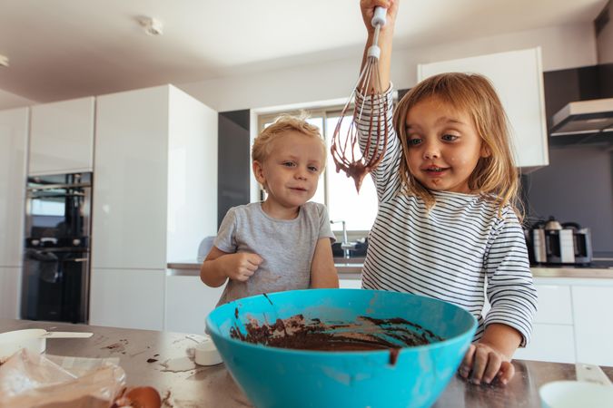 Little girl  whipping chocolate cream with whisk in bowl with brother supervising in kitchen