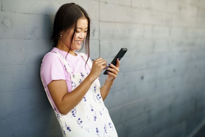 Happy Asian female wearing casual clothes holding phone while leaning against wall, copy space