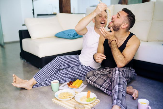 Male couple eating pasta together at home sitting on floor