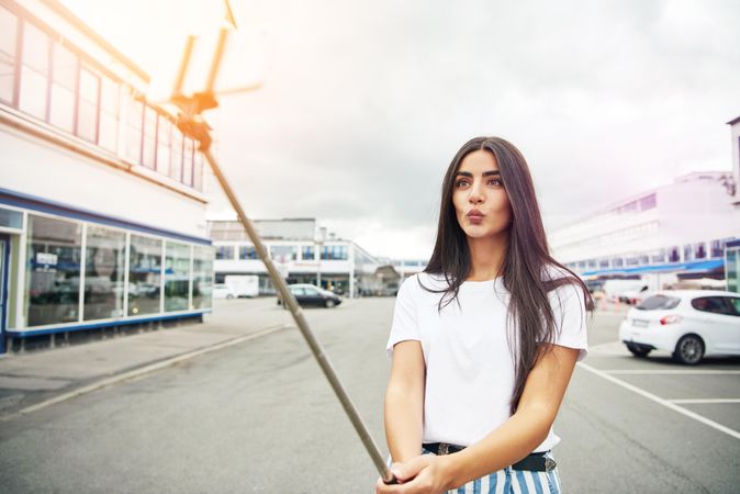 Woman pouting with selfie stick on street with parked cars