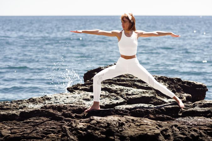 Woman wearing sport clothes and doing warrior pose on rocks on the coast