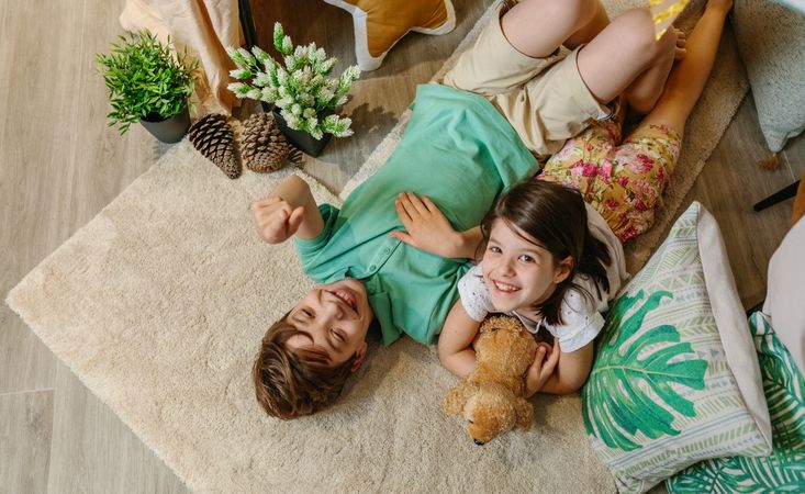 Children laughing and relaxing lying over rug in cozy tent at home surrounded by cushions and plants