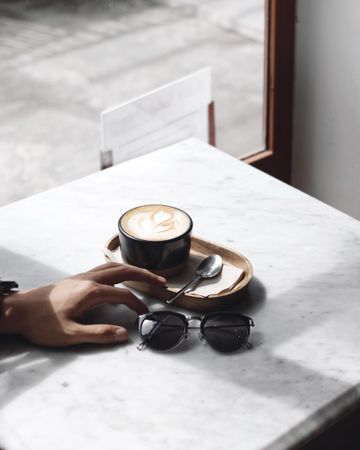 Person's hand on table beside a cup cappuccino and sunglasses during the day