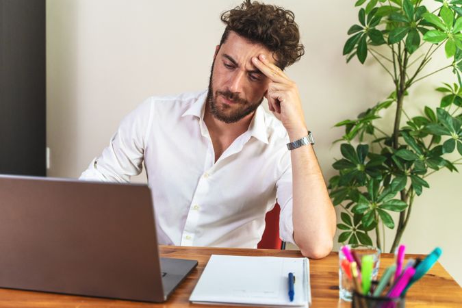Stressed young man with a headache working on his laptop at a desk.