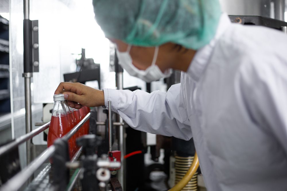 Asian man worker inspecting drink product on bottling factory machinery.