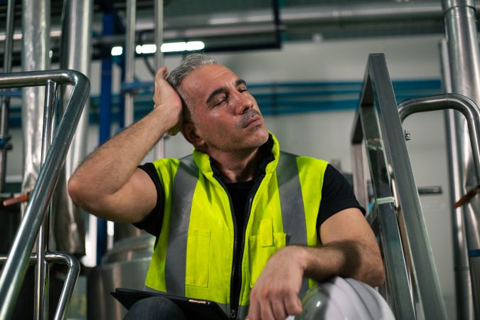 A male industrial engineer is sitting down and resting with his hand on his forehead, showing signs of fatigue or stress after long hours of work in a factory environment.