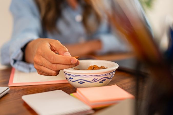 Close-up of a woman’s hand picking almonds from a bowl on a desk with notebooks