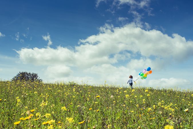 Unrecognizable child walking in grassy countryside with balloons